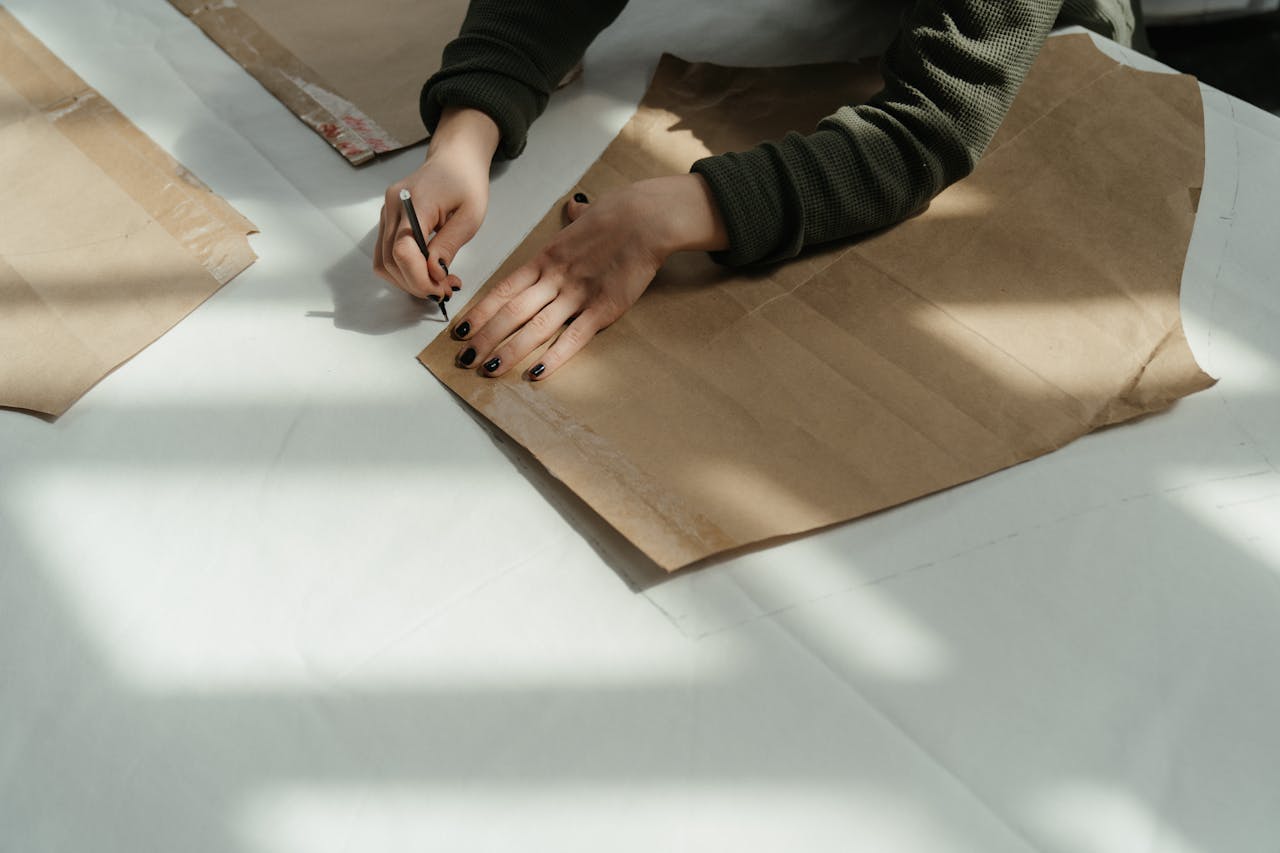 about-me-img Tailor tracing patterns on brown paper in a sunlit workshop, capturing the art of fashion design.